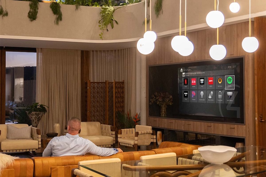 Man on sofa controlling Control4 touchscreen with lighting and streaming apps in a luxury living room.